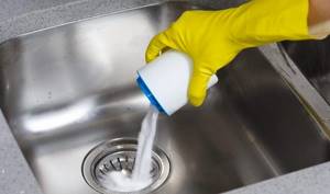 A gloved hand pours powder into the sink drain